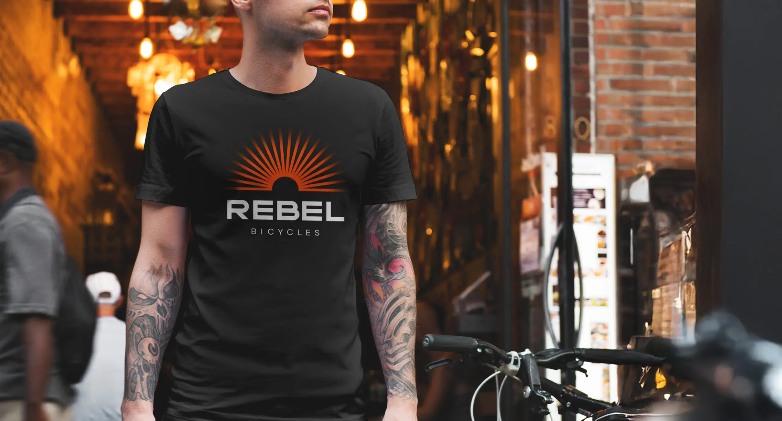 Фірмова чорна футболка з логотипом Rebel Bicycles на хлопці в татуюваннях біля велосипеду.