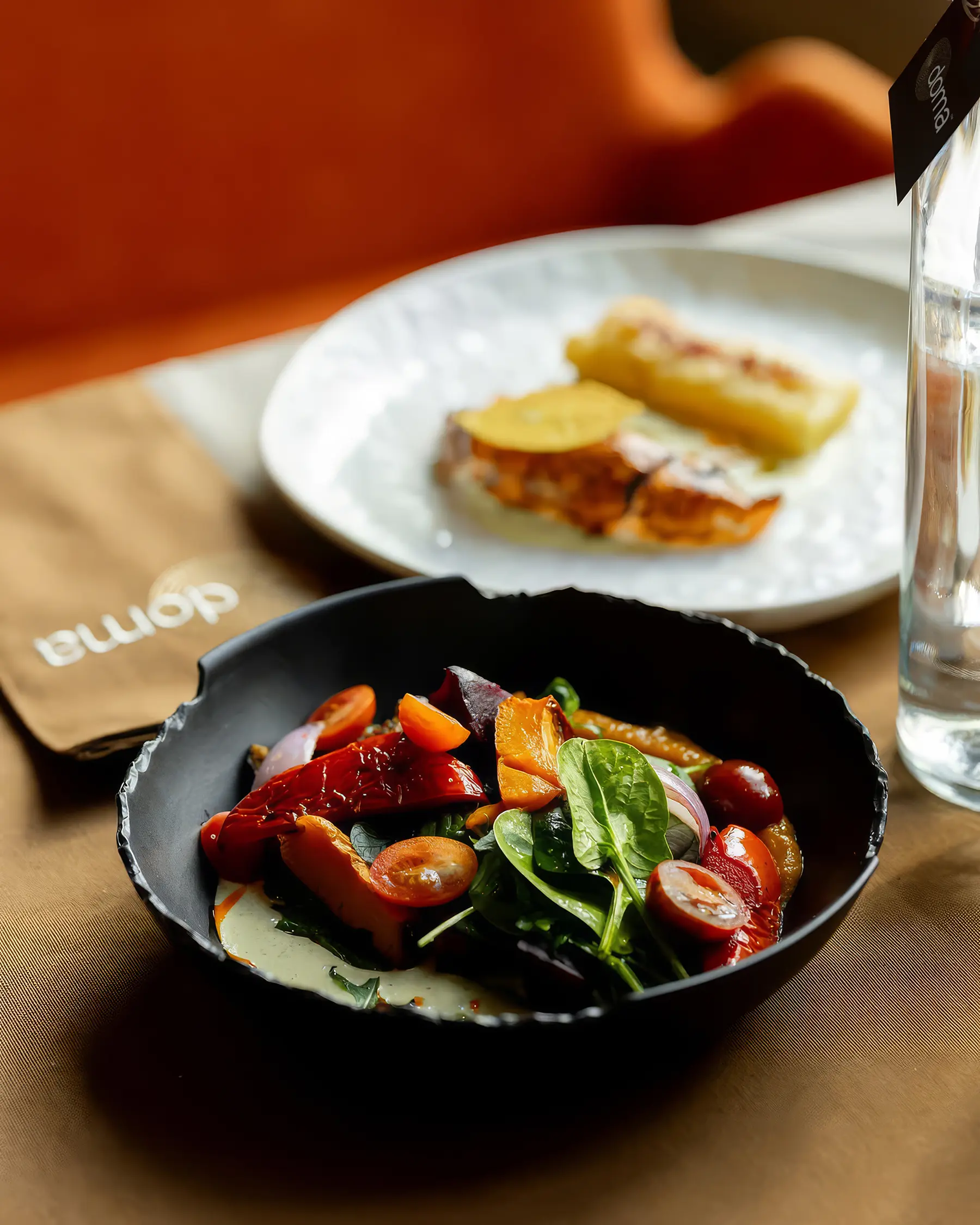 Салат у чорному керамічному посуді поруч з десертом і брендованою серветкою ресторану doma.